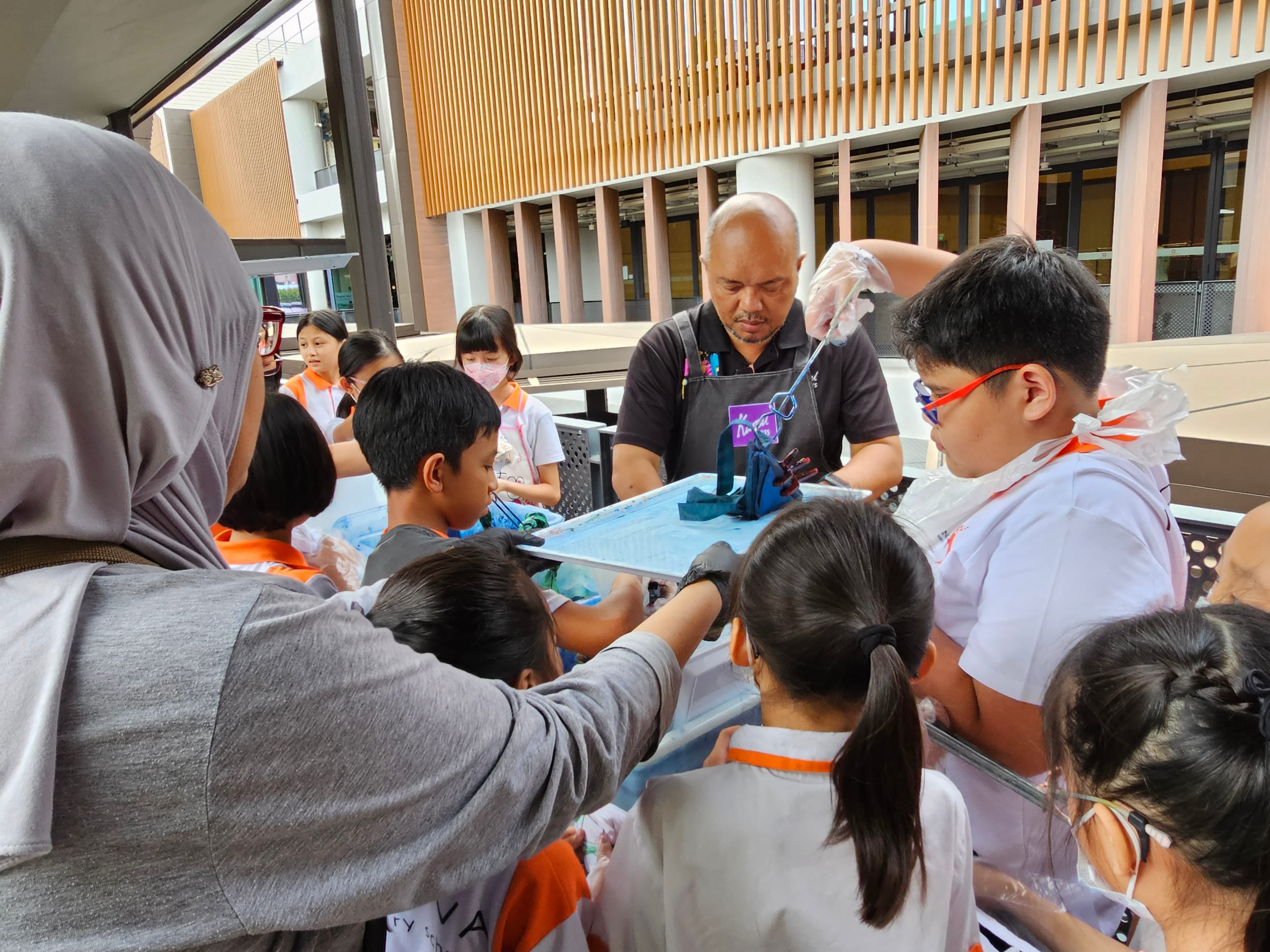 An adult instructor wearing an apron guides a group of children in a hands-on craft activity outdoors.